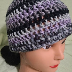 May include: A handmade crochet beanie in shades of gray, black, and lavender. The hat is made with a textured stitch, creating a visually interesting pattern. The beanie is shown on a mannequin head.