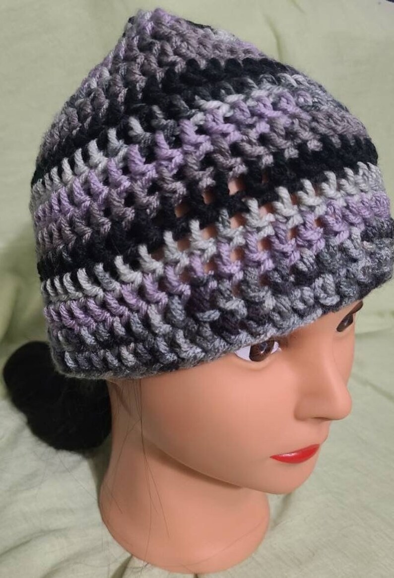 May include: A handmade crochet beanie in shades of gray, black, and lavender. The hat is made with a textured stitch, creating a visually interesting pattern. The beanie is shown on a mannequin head.