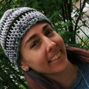 May include: A crocheted beanie hat in shades of gray, black, and lavender. The hat has a textured, open weave design. The hat is worn by a person with long, reddish-purple hair.