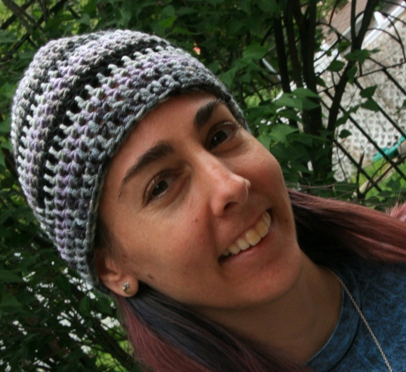 May include: A crocheted beanie hat in shades of gray, black, and lavender. The hat has a textured, open weave design. The hat is worn by a person with long, reddish-purple hair.