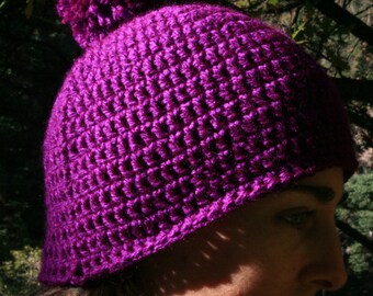 May include: A purple crocheted beanie with a pom pom on top. The beanie is worn by a person, and the person's face is visible.
