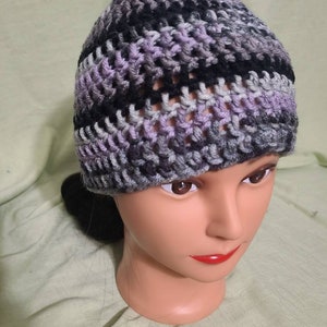 May include: A handmade crochet beanie in a mix of colors including lavender, black, gray, and white. The hat is made with a textured stitch, and is displayed on a mannequin head. The beanie is a winter accessory.