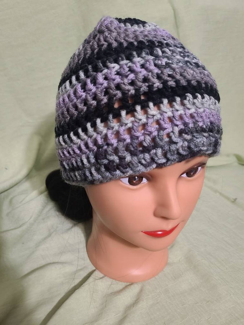 May include: A handmade crochet beanie in a mix of colors including lavender, black, gray, and white. The hat is made with a textured stitch, and is displayed on a mannequin head. The beanie is a winter accessory.