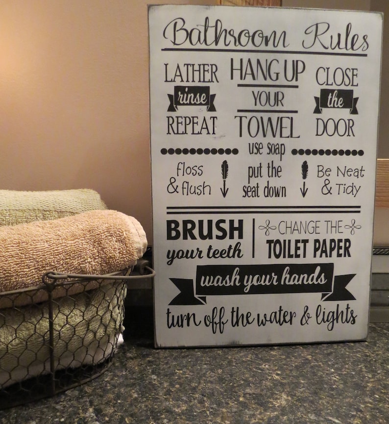 May include: A rectangular white wooden sign with black text reading Bathroom Rules, Lather rinse repeat, Hang up your towel, Close the door, use soap, floss & flush, put the seat down, Be Neat & Tidy, Brush your teeth, Change the toilet paper, wash your hands, turn off the water & lights.