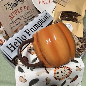 May include: A pumpkin-shaped ceramic mug with a brown handle, a wooden sign reading Homemade Apple Pie, a white spoon rest with Hello Fall text, a brown paper bag with pinecones, and a white cloth featuring a pattern of mushrooms and leaves.
