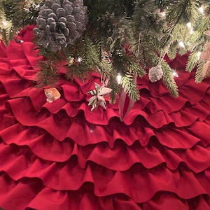 May include: A red ruffled Christmas tree skirt with multiple layers of fabric. The skirt is made of a soft, plush material and has a classic, elegant design.