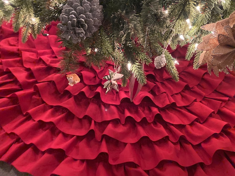 May include: A red ruffled Christmas tree skirt with multiple layers of fabric. The skirt is made of a soft, plush material and has a classic, elegant design.