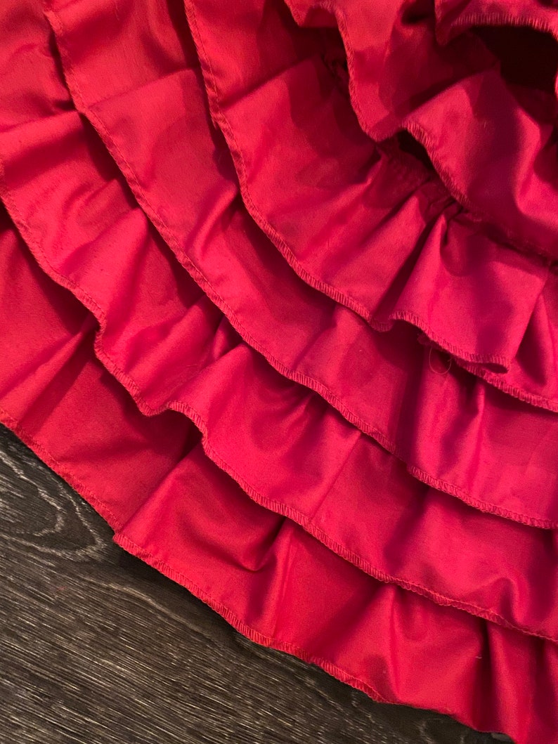 May include: A close-up of a red fabric skirt with multiple layers of ruffles. The ruffles are sewn with a zig-zag stitch.