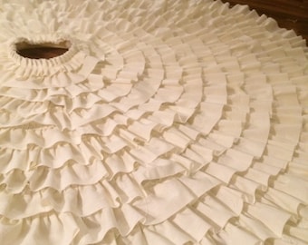 May include: A white, circular, fabric skirt with multiple layers of ruffles. The skirt is made of a soft, lightweight fabric and has a gathered waistband.
