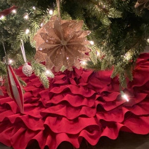May include: A red ruffled Christmas tree skirt with multiple layers of fabric. The skirt is placed under a Christmas tree with lights and ornaments.