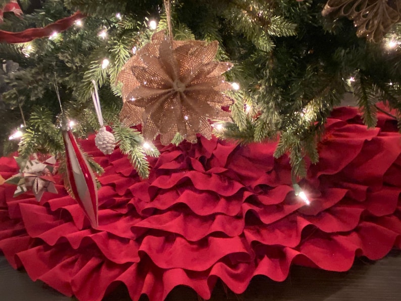 May include: A red ruffled Christmas tree skirt with multiple layers of fabric. The skirt is placed under a Christmas tree with lights and ornaments.