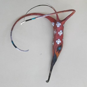 May include: An abstract sculpture with a red body decorated with white flower details. The sculpture has a curved, elongated form with a black and silver tail and a curved arm. The artwork features a mix of colours, including red, black, silver, and blue.