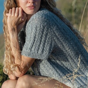 Puede incluir: Una mujer con el pelo largo y rizado rubio se sienta en un campo de hierba alta. Lleva un jersey de punto gris y pantalones cortos blancos. El fondo es un cielo azul y una masa de agua.