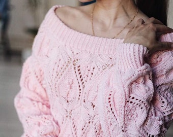 May include: A pink knitted sweater with a loose fit and an open-stitch design. The sweater has a wide neckline and long sleeves.