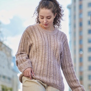 Puede incluir: Una mujer con un suéter de punto de cable marrón claro con un patrón de costillas. Lleva pantalones beige y tiene el pelo recogido en una coleta.