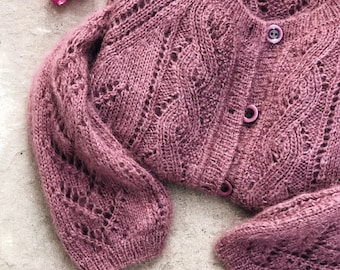 May include: A mauve knitted cardigan with a delicate lace pattern and button closures. The cardigan is laid out on a textured surface with pink flowers.