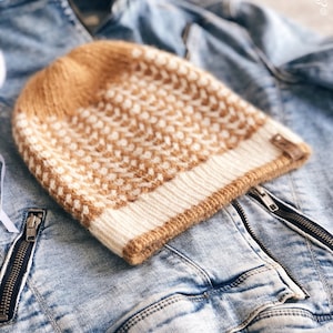 May include: A brown and white knitted beanie with a geometric pattern. The beanie is sitting on a blue denim jacket.