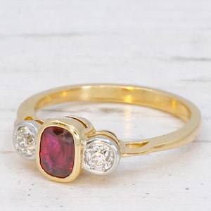 May include: A gold ring featuring a central cushion-cut red ruby gemstone set in a yellow gold bezel, flanked by two round-cut white diamonds, each secured in a silver-toned metal setting, creating a three-stone design on a polished gold band.