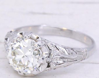 May include: A silver engagement ring with a large, round, clear diamond. The ring features intricate floral engravings on the band and around the diamond setting. The ring is set against a white background.