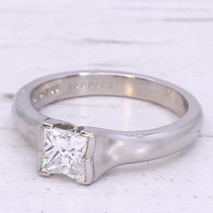 Può includere: Un anello di fidanzamento in platino con un diamante di forma quadrata incastonato in una fascia semplice. L'anello è inciso con le parole "Leo Palais".
