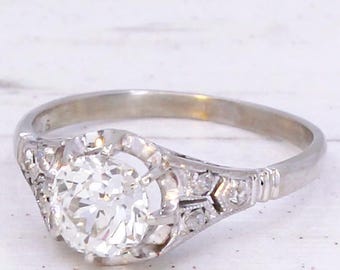 May include: A vintage-style engagement ring with a large, round, clear diamond as the centerpiece. The ring has a silver band with intricate detailing on the sides, and small accent diamonds. The ring is set against a white background.