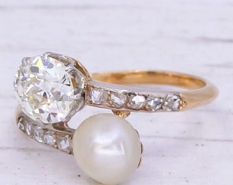 May include: An antique gold ring featuring a large, sparkling diamond and a dangling pearl. The band is accented with smaller diamonds. The ring's design is elegant and vintage, showcasing a classic jewelry style.