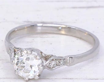 May include: A silver engagement ring with a large, round, clear gemstone as the centerpiece. The band has a delicate design with smaller accent stones. The ring is set against a white background.