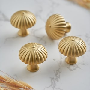 May include: Four gold-colored cabinet knobs with a mushroom-like design. Each knob features a ribbed, semi-circular top and a short, rounded base. The knobs are arranged on a white marble surface, with a few sprigs of dried grass in the background.