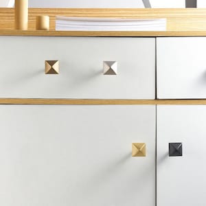 Op de afbeelding: Vier piramidevormige kastknoppen in goud, zilver, messing en zwart. De knoppen zijn gemonteerd op een witte kastdeur.