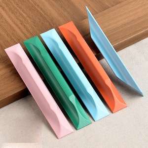 Op de afbeelding: Zes kastgrepen in verschillende kleuren, waaronder roze, groen, blauw en oranje. De grepen zijn gemaakt van metaal en hebben een gebogen ontwerp.