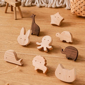 Peut inclure: Diverses figurines d'animaux en bois, dont une girafe, un lapin, une pieuvre, une tortue, un éléphant, une baleine, un lion et un chat. Les figurines sont marron clair et foncé, avec des designs minimalistes. Un petit cerf en bois et un panier sont en arrière-plan.
