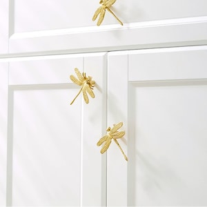 Könnte beinhalten: Drei goldene Schrankknöpfe in Libellenform, jeweils mit filigranen Details, sind an einer weißen Schranktür montiert.