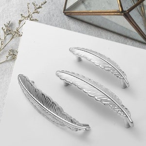 Può includere: Tre maniglie per armadi a forma di piuma argentate. Le maniglie sono tutte dello stesso design, ma di dimensioni diverse. Le maniglie hanno un design a piuma dettagliato.