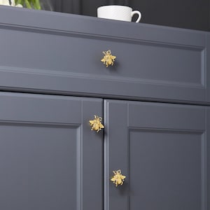 May include: Three gold bee-shaped cabinet knobs on a grey cabinet. The knobs are detailed with wings and legs.