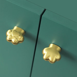 May include: Two gold paw-shaped cabinet knobs on a teal green cabinet door.