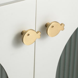 May include: Two gold fish-shaped cabinet knobs with a simple eye design. The knobs are mounted on a white cabinet door.