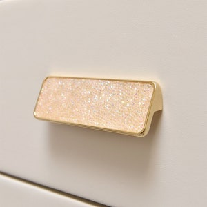 Puede incluir: Un tirador de cajón dorado con forma rectangular y una superficie brillante e iridiscente.