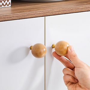 May include: Two round wooden knobs with gold metal hardware. The knobs are mounted on a white cabinet door.