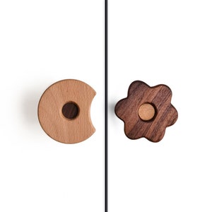 以下が含まれることがあります： 木製のキャビネットノブ2個、1つは切り抜きのある丸型、もう1つは花の形をしています。丸いノブは明るい茶色の木で、花のノブは濃い茶色の木です。