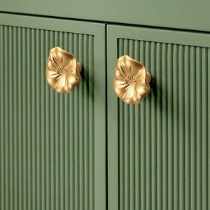 May include: Close-up of a green cabinet with vertical ribbed detailing. The cabinet doors feature two gold-coloured, flower-shaped knobs. The knobs are detailed with petals and a central floral design.