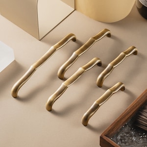 May include: Five gold-toned cabinet pulls with a bamboo-inspired design. The pulls are arranged in a staggered pattern on a beige surface.