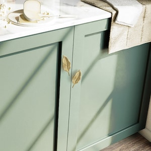 May include: Two gold leaf-shaped cabinet pulls are mounted on a pair of sage green cabinet doors. The cabinet doors are closed and the cabinet is in a kitchen setting.
