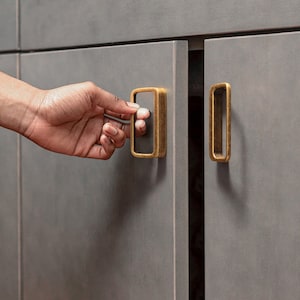 Op de afbeelding: Close-up van een kastdeur met een rechthoekige, antiek messingkleurige handgreep. De handgreep wordt opengemaakt. De kastdeur heeft een effen, matte grijze kleur.