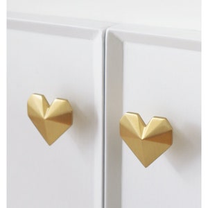 May include: Two gold-colored, geometric heart-shaped cabinet knobs are mounted on a white cabinet. The knobs have a faceted design, catching the light and adding a touch of elegance to the cabinet doors.