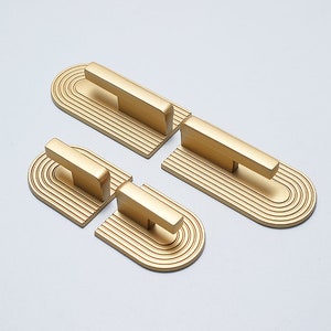 Op de afbeelding: Set van vier gouden kastgrepen met een afgeronde rechthoekige vorm en een verhoogd, gegroefd ontwerp. Elke greep heeft een eenvoudige, horizontale staaf.