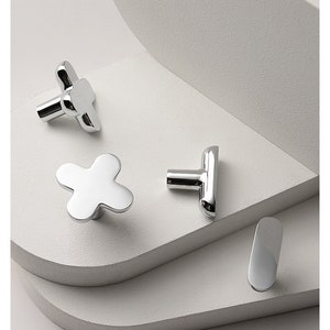 以下が含まれることがあります： 4つの異なる形のシルバーのキャビネットノブ。ノブは白い表面にあります。