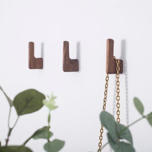 May include: Three wooden wall hooks in a light brown colour. The hooks are shaped like an L and are mounted on a white wall. A gold chain is hanging from one of the hooks.