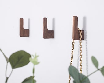 May include: Three wooden wall hooks in a light brown color. The hooks are shaped like an L and are mounted on a white wall. A gold chain is hanging from one of the hooks.