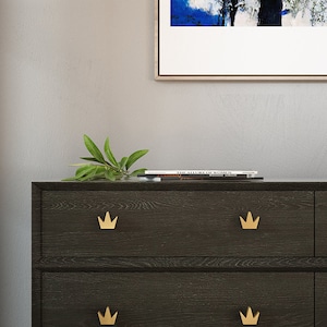 May include: A dark wood dresser with four drawers. Each drawer has a gold crown-shaped pull. The dresser is topped with a book titled "The Nature of Women".