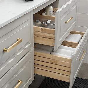Puede incluir: Primer plano de un tocador de baño con tres cajones. El cajón superior está abierto, revelando un organizador de cajones de madera con dos botellas y un plato pequeño. El cajón del medio está cerrado y tiene una manija de color dorado. El cajón inferior está abierto y tiene una manija de color dorado. El tocador está pintado de color gris claro.